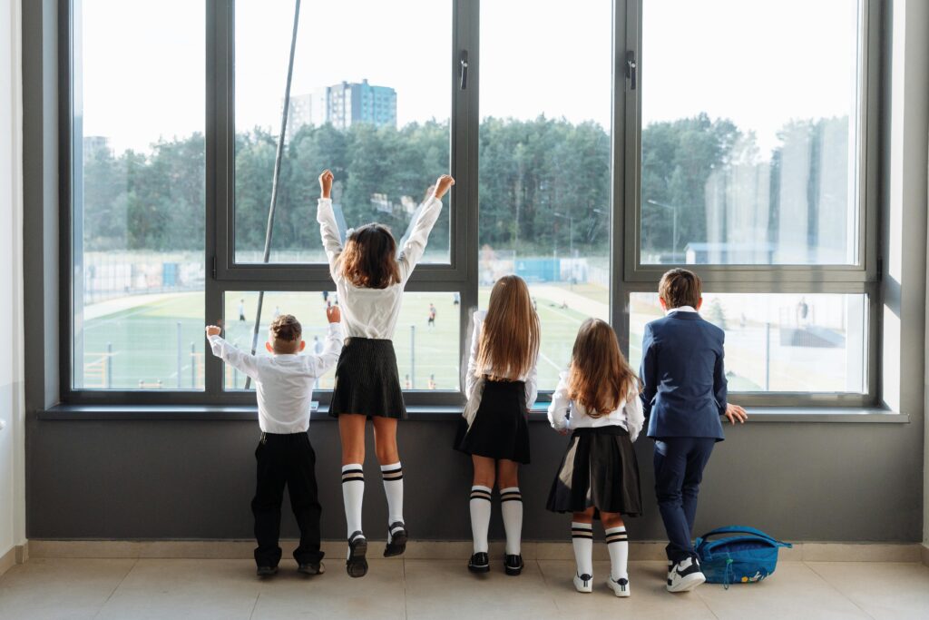 A group of school children in uniform stand by a large window, looking outside.